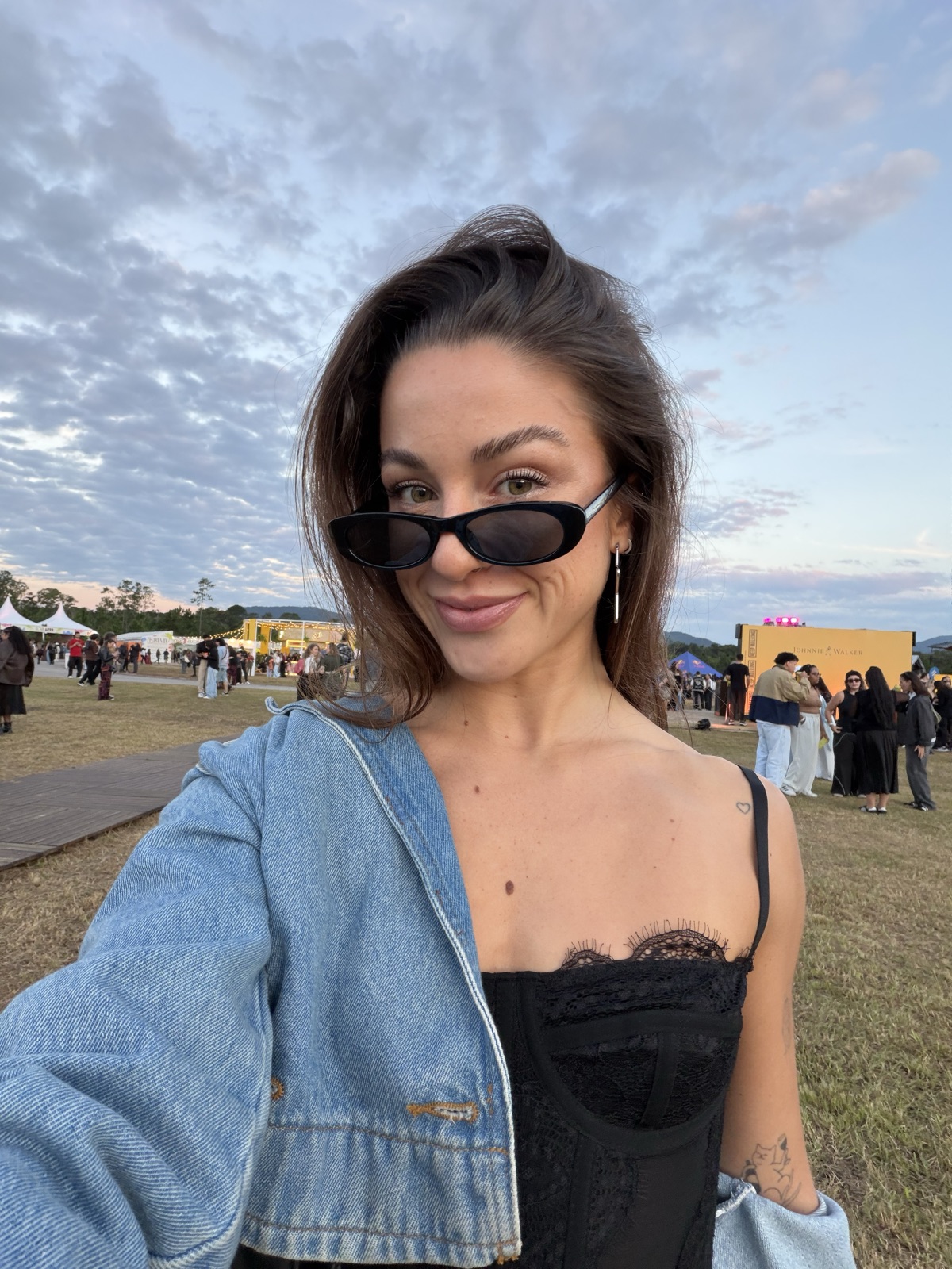 Gabi de óculos escuros e jaqueta jeans, em um festival ao ar livre no fim da tarde