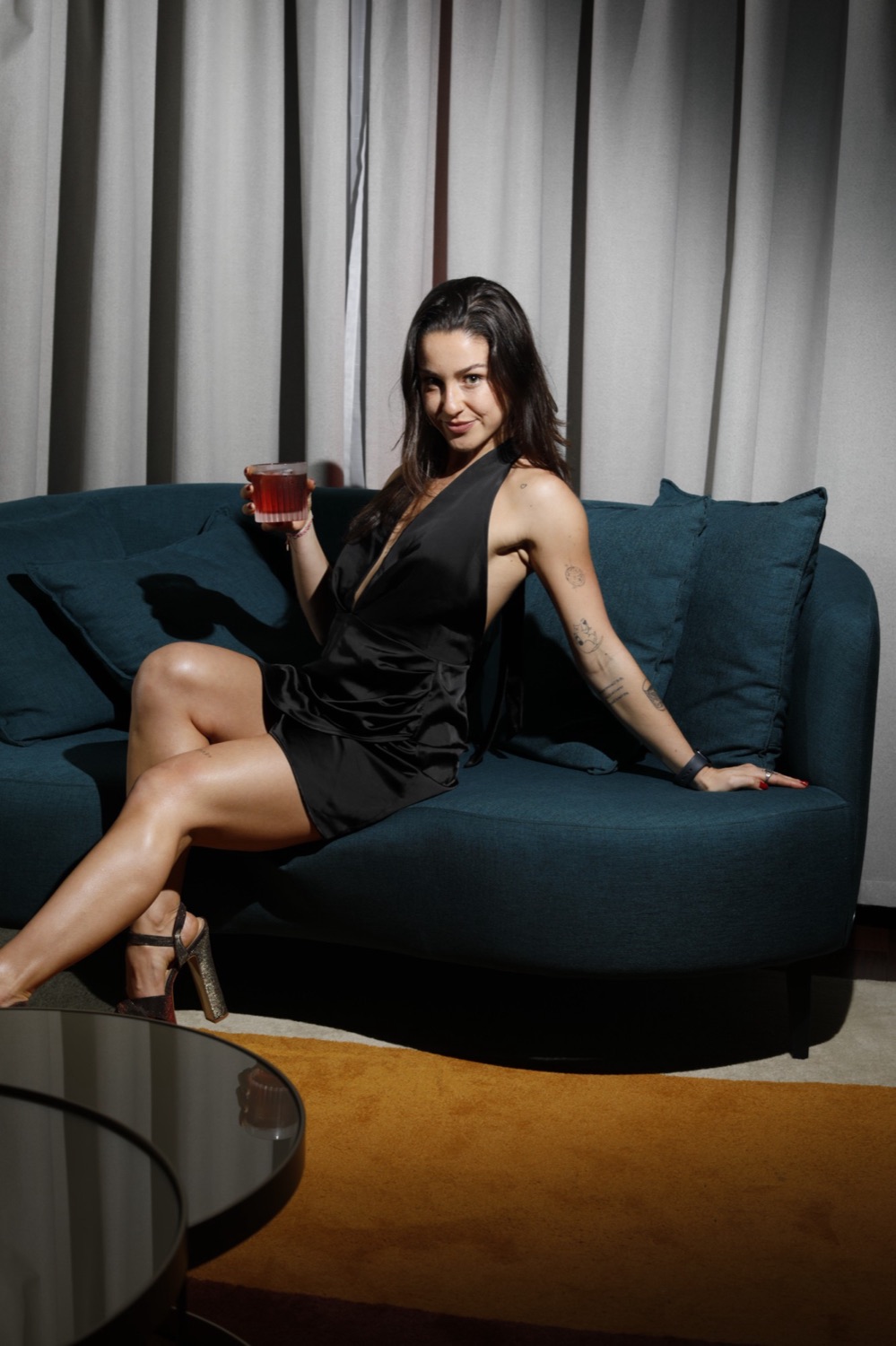 Gabi em um sofá azul, de vestido preto, com um drink na mão